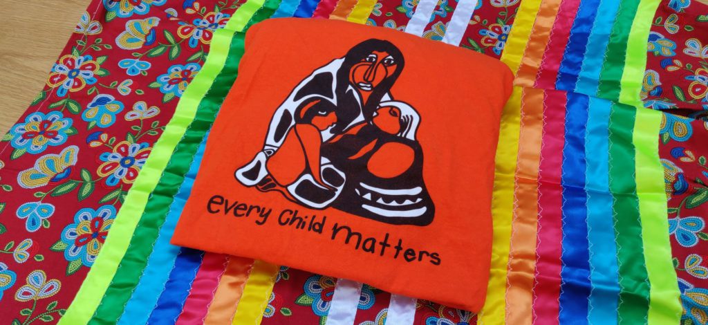 Image of an orange t-shirt with "every child matters" written on it in black text. Behind is a ribbon skirt.
